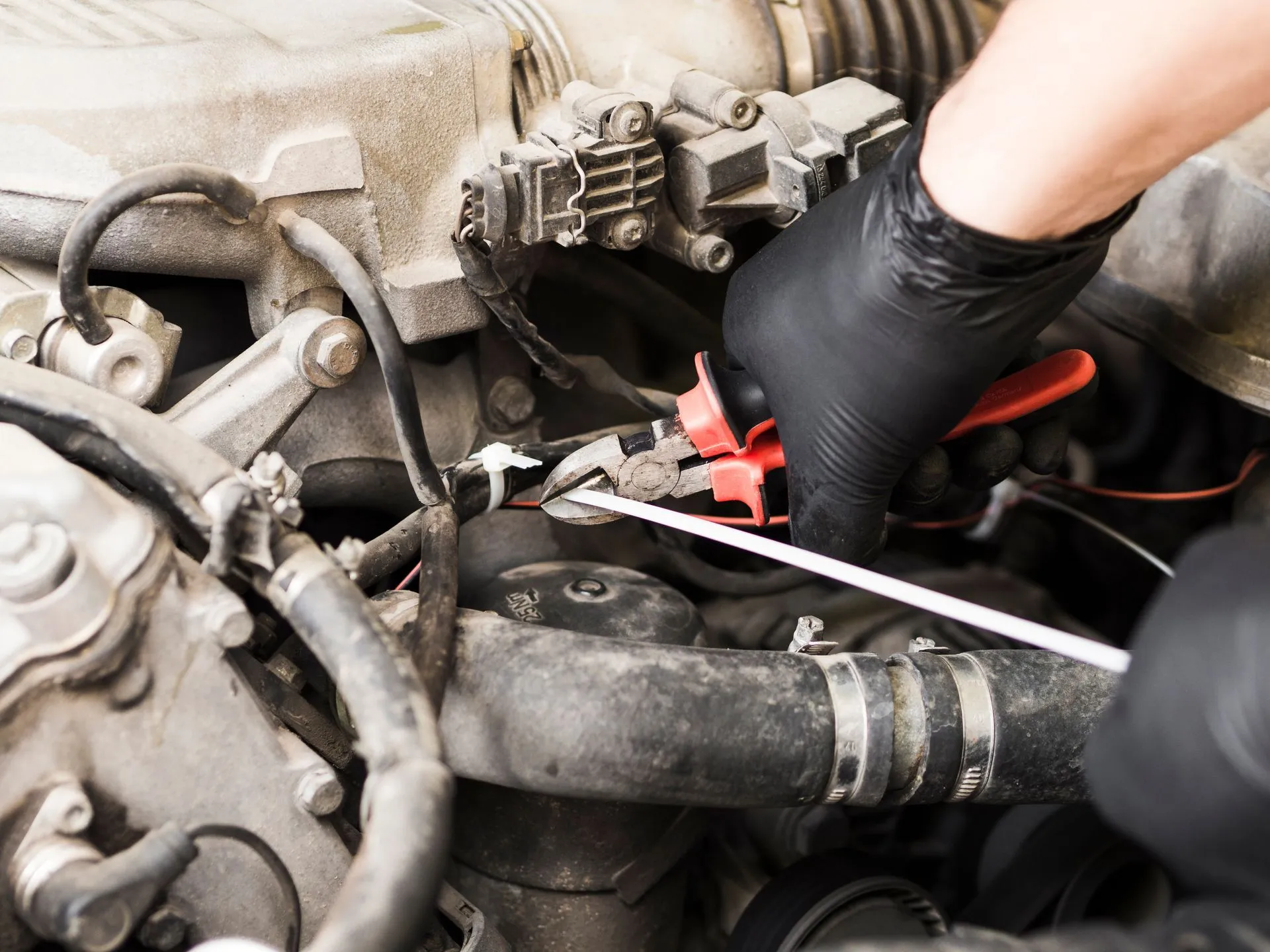 Gloved technician cutting a cable tie on engine bay wiring with red-handled side cutters